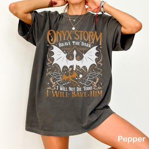 May include: Dark gray t-shirt with a graphic design featuring the text "ONYX STORM BRAVE THE DARK" and the phrases "I WILL NOT DIE TODAY I WILL SAVE HIM." The design includes a bat and other fantasy elements.