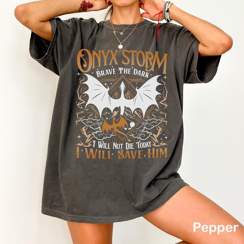 May include: Dark gray t-shirt with a graphic design featuring the text "ONYX STORM BRAVE THE DARK" and the phrases "I WILL NOT DIE TODAY I WILL SAVE HIM." The design includes a bat and other fantasy elements.