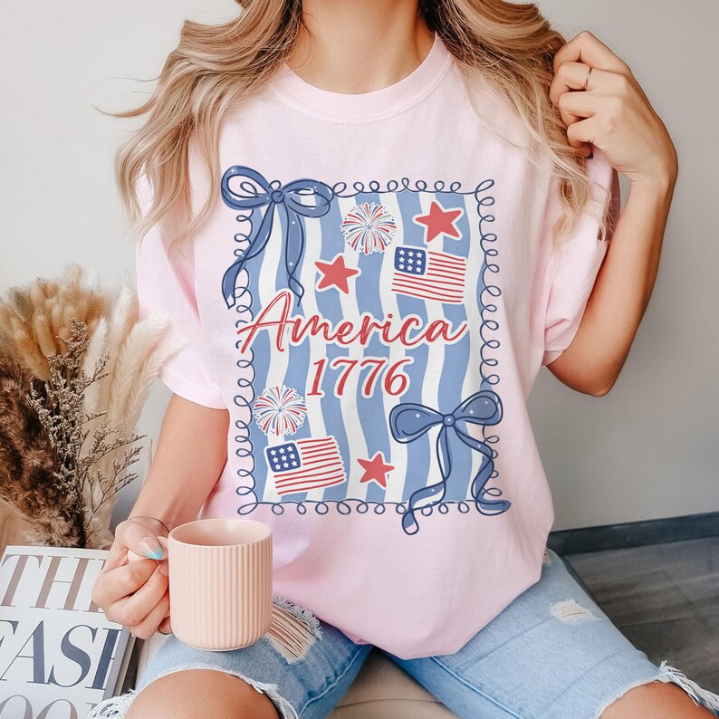 May include: A pale pink t-shirt with a patriotic design. The design features the text "America 1776" in red script, along with blue bows, red stars, and American flags. The shirt has a relaxed fit.