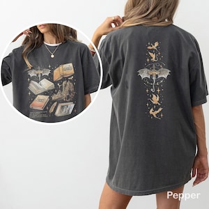 May include: Dark grey t-shirt with a fantasy graphic. The front displays a dragon, books, and castle illustrations. The back has a dragon and other flying creatures. The shirt has a relaxed fit.