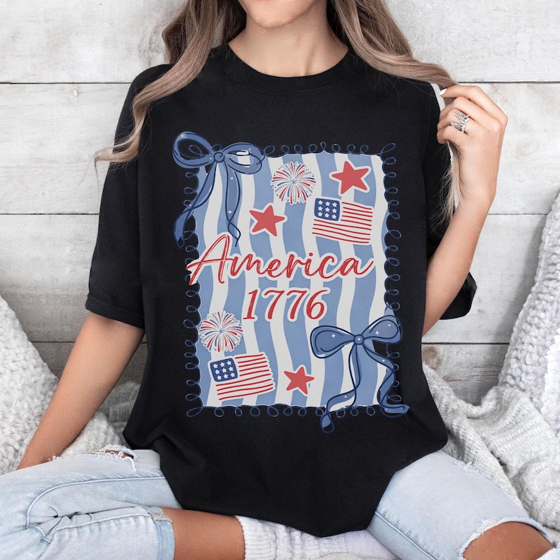 May include: Black t-shirt with a patriotic design. The design features the text "America 1776", red stars, blue bows, fireworks, and American flags on a blue and white striped background.