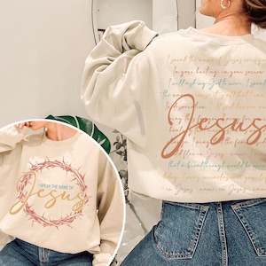 May include: Beige sweatshirt with the text "I speak the name of Jesus" in a gold script, surrounded by a crown of thorns. The back of the sweatshirt has a script with the word "Jesus" in a large, stylized font.
