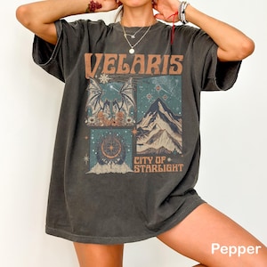 Puede incluir: Camiseta gris oscuro con un diseño gráfico de cuatro paneles. El panel superior dice "VELARIS" en letras naranjas. Los otros paneles presentan ilustraciones de un murciélago, montañas y una luna creciente. El texto "CITY OF STARLIGHT" está en la parte inferior.