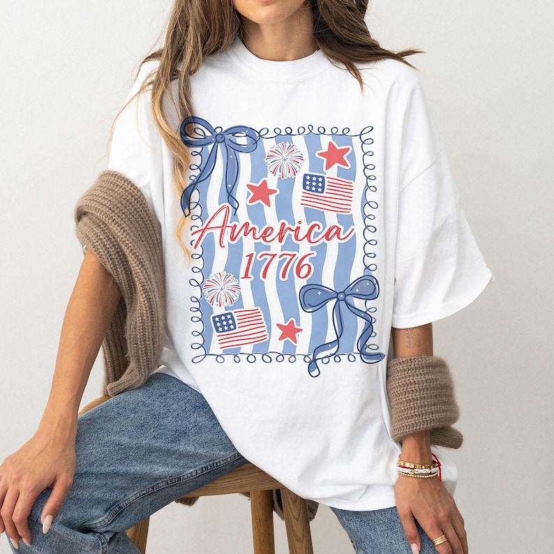 May include: White t-shirt featuring a patriotic design. The design includes the words "America 1776" in red script, blue bows, red stars, and American flags. The background has blue and white vertical stripes.