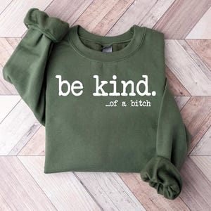 Puede incluir: Sudadera verde oliva con la frase "be kind. ...of a bitch" en blanco. La sudadera tiene cuello redondo y mangas largas con puños fruncidos. La prenda está sobre una superficie clara con vetas de madera.