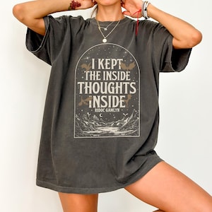 Op de afbeelding: Donkergrijs t-shirt met een witte afbeelding met de tekst "I KEPT THE INSIDE THOUGHTS INSIDE" in een gestileerde boog. Het ontwerp bevat vleermuizen, bergen en de naam van de auteur, Ridoc Gamlyn. Het shirt heeft korte mouwen en een relaxte pasvorm.