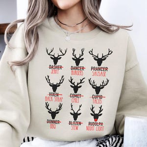 May include: A cream-colored sweatshirt featuring black reindeer silhouettes with humorous food-related names like "Jerky" and "Burgers" underneath. The word "Rudolph" has a red nose. The sweatshirt is a festive and playful design.