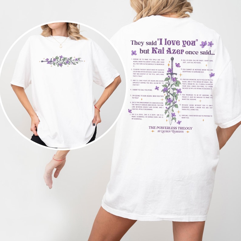 Puede incluir: Camiseta blanca con un dise&ntilde;o floral y de espada en la parte delantera y texto en la parte trasera. La parte trasera de la camiseta tiene el texto "They said 'I love you' but Kai Azer once said..." y una lista de citas. La camiseta es parte de "The Powerless Trilogy".