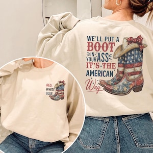 May include: A cream-colored sweatshirt featuring a patriotic design. The front displays a small graphic of a cowboy boot in red, white, and blue with the words "Red, White, Blue." The back has a larger graphic with text and a cowboy boot.