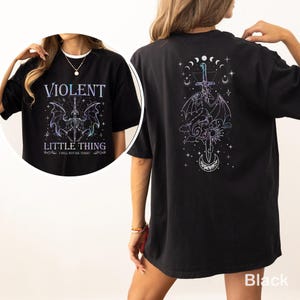 Op de afbeelding: Zwart T-shirt met de tekst "VIOLENT LITTLE THING" en een vleermuisachtig wezenontwerp op de voorkant. De achterkant toont een draak- en zwaardontwerp met hemelse elementen. Het shirt heeft een relaxte pasvorm.