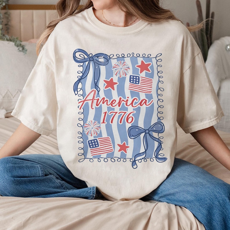 May include: A cream-coloured t-shirt with a patriotic design. The graphic features the word "America" in red script, the year "1776", red stars, blue bows, and American flags. The design is framed by a blue border.