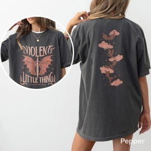 May include: Dark gray t-shirt with the text "VIOLENT LITTLE THING" and a graphic of a sword with wings on the front. The back of the shirt features a design of clouds and bats. The shirt is a Pepper color.
