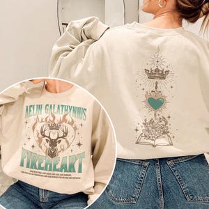 May include: Cream-coloured sweatshirt with a graphic design. The front features text that reads "AELIN GALATHYNIUS FIREHEART" with a deer, crown, and flames. The back has a crown, sword, heart, and book design. The text "HE WAS FIRE AND LIGHT AND ASH AND EMBERS" is also visible.