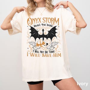 May include: Ivory-colored t-shirt featuring a black bat graphic with the text "ONYX STORM BRAVE THE DARK" and "I WILL NOT DIE TODAY I WILL SAVE HIM". The design includes a dragon and decorative elements.