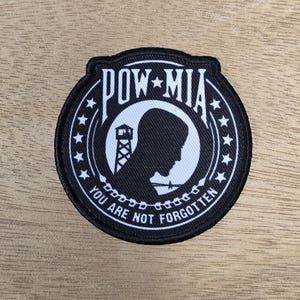 Op de afbeelding: Een zwart-witte POW-MIA-patch. De patch is rond met een zwarte rand en een wit interieur. De tekst "POW-MIA" staat bovenaan en "YOU ARE NOT FORGOTTEN" onderaan. Een silhouet van een persoon en een wachttoren staat in het midden.