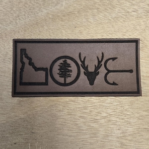 May include: A rectangular brown leather patch with the word "LOVE" spelled out with symbols. The symbols include the outline of the state of Idaho, a tree inside a circle, a deer head, and two fishing hooks.