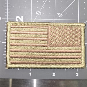 Może przedstawiać: Naszywka z haftowanej amerykańskiej flagi w kolorze beżowym z brązowym obramowaniem. Flaga wykonana jest z miękkiej, fakturowanej tkaniny.