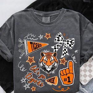 May include: Dark gray t-shirt with a tiger head and the words "Tigers" and "Let's go tigers" in orange and white. The shirt also features a checkered bow, stars, and a #1 hand symbol.