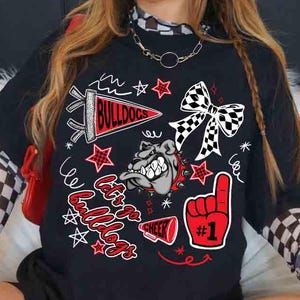 May include: Black t-shirt with a graphic design featuring the word "Bulldogs", a bulldog mascot, stars, and a foam finger with the number one. The design includes red, white, and black elements. The shirt has short sleeves.