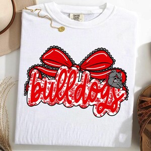 May include: White t-shirt featuring the word "Bulldogs" in red script with a floral pattern, topped with a red bow. A gray bulldog graphic is on the right. This is a piece of clothing.