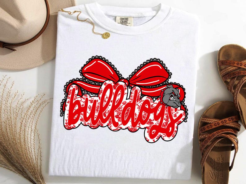 May include: White t-shirt featuring the word "Bulldogs" in red script with a floral pattern, topped with a red bow. A gray bulldog graphic is on the right. This is a piece of clothing.