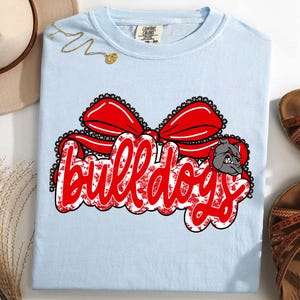 May include: Pale blue t-shirt featuring the word "Bulldogs" in red script, with a red bow and a bulldog graphic. The text has a floral pattern. The shirt is a Comfort Colors brand.