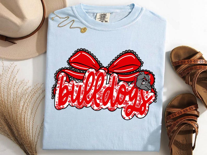 May include: Pale blue t-shirt featuring the word "Bulldogs" in red script, with a red bow and a bulldog graphic. The text has a floral pattern. The shirt is a Comfort Colors brand.