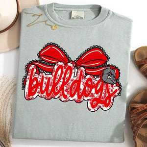 May include: A pale blue t-shirt featuring a red bow design and the word "Bulldogs" in a decorative font. A bulldog graphic is on the right. The shirt is displayed with sandals and a hat.
