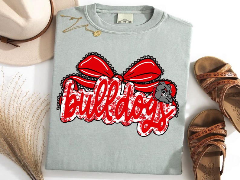 May include: A pale blue t-shirt featuring a red bow design and the word "Bulldogs" in a decorative font. A bulldog graphic is on the right. The shirt is displayed with sandals and a hat.