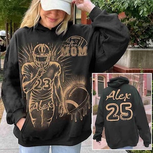 Puede incluir: Sudadera con capucha negra con un diseño de fútbol. La parte delantera presenta un gráfico de jugador de fútbol, el número 23 y el nombre "Alex". También es visible el texto "Football Mom". La parte trasera de la sudadera tiene el nombre "Alex" y el número 25.