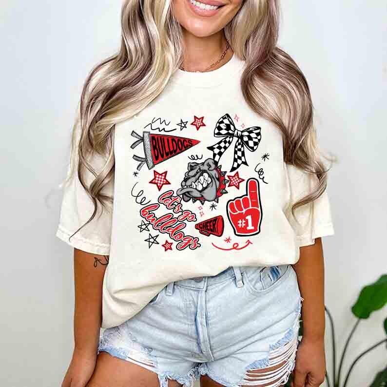 May include: Cream-colored t-shirt featuring a graphic design with the text "Let's Go Bulldogs". The design includes a bulldog, stars, a bow, and a foam finger. The shirt is a comfortable, casual style.