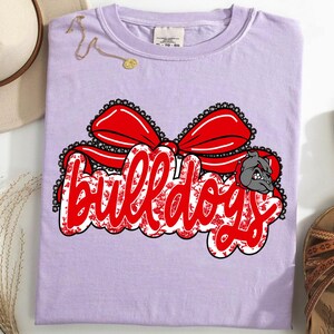 May include: A lilac t-shirt featuring the word "Bulldogs" in red script with a floral design, a red bow, and a bulldog graphic. The shirt is accompanied by a hat and sandals.