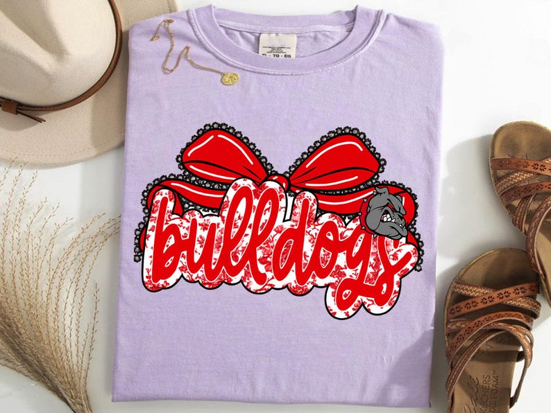 May include: A lilac t-shirt featuring the word "Bulldogs" in red script with a floral design, a red bow, and a bulldog graphic. The shirt is accompanied by a hat and sandals.