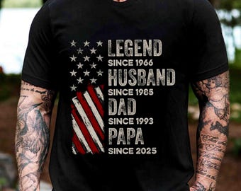 May include: Black t-shirt featuring an American flag design with the text "LEGEND SINCE 1966, HUSBAND SINCE 1985, DAD SINCE 1993, PAPA SINCE 2025." The design is distressed, giving it a vintage look. The shirt is a classic crew neck.