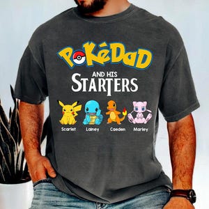 May include: Dark gray t-shirt featuring the text "PokéDad and His Starters" in yellow and white. Below the text are four cartoon characters: Pikachu, Squirtle, Charmander, and Mew. The names Scarlet, Lainey, Caeden, and Marley are below each character.