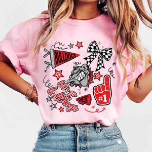 May include: Pink t-shirt with a graphic design featuring a bulldog, stars, a bow, and the words "Let's Go Bulldogs". The design includes a red foam finger with the number one.