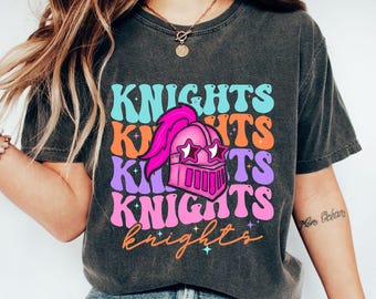 May include: Dark gray t-shirt with the word "KNIGHTS" in a retro font, stacked in multiple colors. A pink knight's helmet with star-shaped eye holes and a feather plume is centered on the shirt. The word "knights" is written in cursive at the bottom.