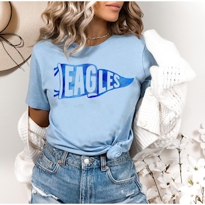 May include: Light blue t-shirt with the word "EAGLES" in a blue pennant design. The shirt is a short-sleeved crew neck style. The model is wearing blue denim shorts and a white cardigan.