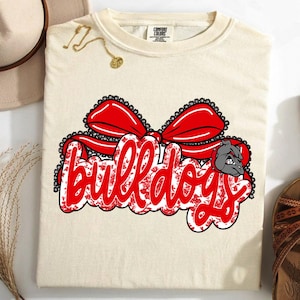 May include: A cream-coloured t-shirt with the word "Bulldogs" in red, with a floral pattern and a red bow design. A grey bulldog graphic is on the right. The shirt is next to a hat, necklace, and sandals.