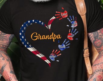 May include: Black t-shirt featuring a heart-shaped design in the colors of the American flag. The word "Grandpa" is in gold script. Handprints with names like "Carter" and "Stella" are arranged around the heart. The text "NAMES & NICK NAME CAN BE CUSTOMIZED" is at the bottom.