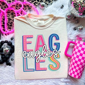 Puede incluir: Camiseta color crema con la palabra "EAGLES" en letras de bloque coloridas y la palabra "eagles" en cursiva. A la derecha hay un vaso a cuadros rosa y blanco con asa rosa.