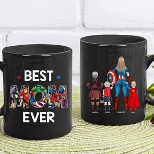 Puede incluir: Dos tazas de cer&aacute;mica negra. Una taza tiene las palabras "BEST MOM EVER" con personajes de superh&eacute;roes en las letras. La otra taza presenta a una mam&aacute; superh&eacute;roe con tres hijos, cada uno con un disfraz de superh&eacute;roe y sus nombres.