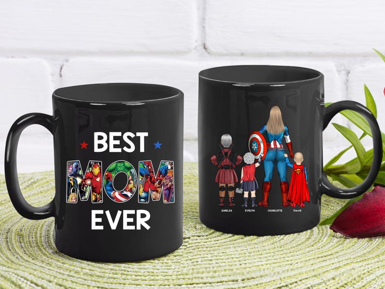 Puede incluir: Dos tazas de cer&aacute;mica negra. Una taza tiene las palabras "BEST MOM EVER" con personajes de superh&eacute;roes en las letras. La otra taza presenta a una mam&aacute; superh&eacute;roe con tres hijos, cada uno con un disfraz de superh&eacute;roe y sus nombres.