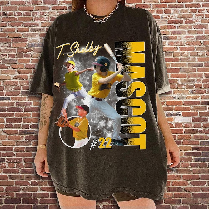 May include: Dark gray t-shirt featuring a baseball-themed design. The shirt displays the name "T. Shelby" in yellow script, along with the word "MASCOT" in large yellow letters. The design includes images of baseball players and the number 22.