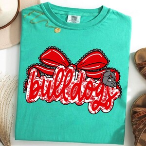 May include: A turquoise t-shirt with a large graphic reading "Bulldogs" in red script with a white floral pattern. A red bow and a bulldog head graphic are above and to the right of the text. The shirt is displayed with sandals and a hat.