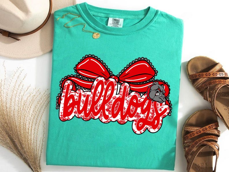 May include: A turquoise t-shirt with a large graphic reading "Bulldogs" in red script with a white floral pattern. A red bow and a bulldog head graphic are above and to the right of the text. The shirt is displayed with sandals and a hat.