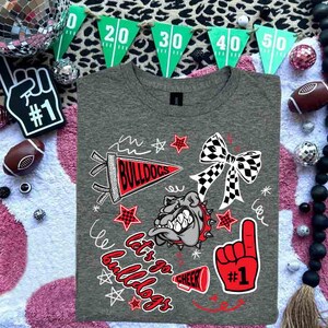 May include: A gray t-shirt with a graphic design featuring a bulldog, pennant, stars, and the words "Let's Go Bulldogs" in red and white. A red foam finger with the number one is also featured. The shirt is surrounded by football-themed decorations.