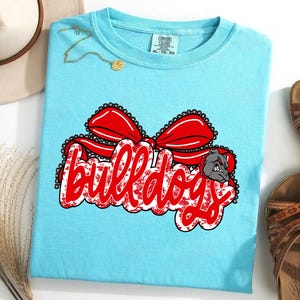 May include: A light blue t-shirt featuring the word "Bulldogs" in red script, a red bow, and a bulldog graphic. This casual t-shirt is suitable for everyday wear.