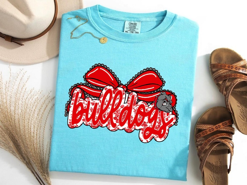 May include: A light blue t-shirt featuring the word "Bulldogs" in red script, a red bow, and a bulldog graphic. This casual t-shirt is suitable for everyday wear.