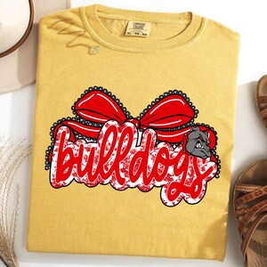 May include: Mustard yellow t-shirt featuring a red bow and the word "bulldogs" in red script. The word "bulldogs" has a floral pattern and a bulldog graphic. A hat and sandals are next to the shirt.